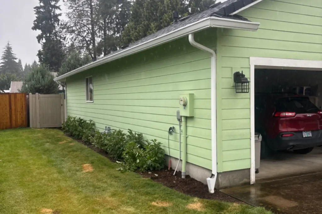 Gutter Installation Scappoose 7 White gutter and downspout installed along the garage side of a light green house in Scappoose, Oregon.