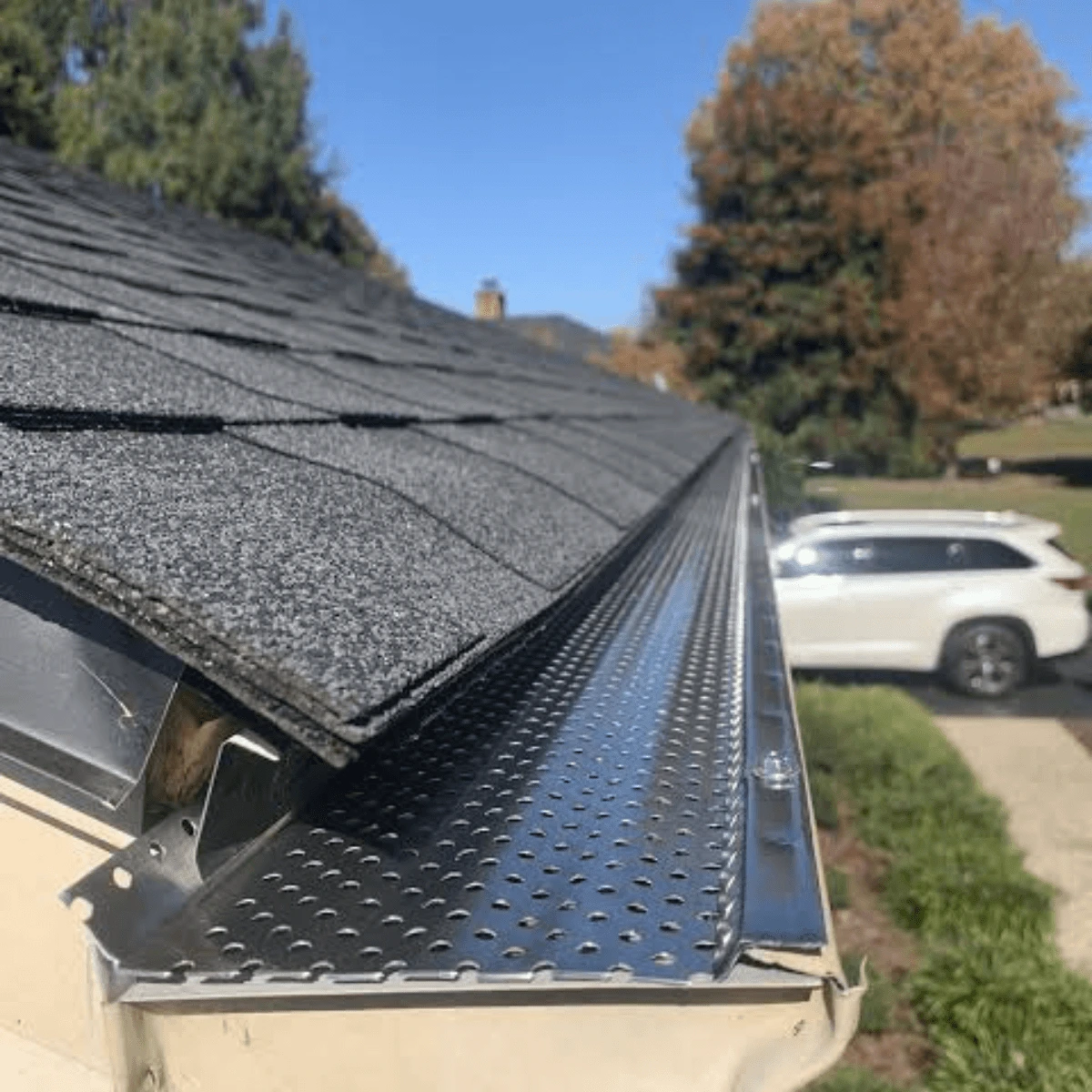 Close-up view of a black perforated gutter guard installed along the edge of an asphalt-shingle roof, with a suburban yard and white SUV in the background.