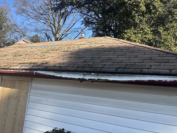 Bent, damaged gutter section and loose trim along the eave of a garage with a mossy shingle roof