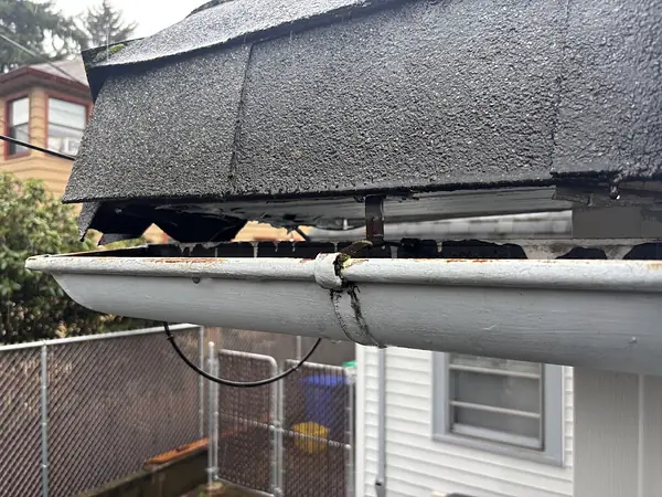 Old gutter hanging on a rusted strap hanger below worn shingles, with the gutter bent at the joint