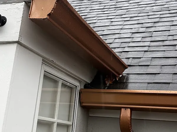 Copper-colored gutters meeting at a roof corner above a white stucco wall, with a matching downspout