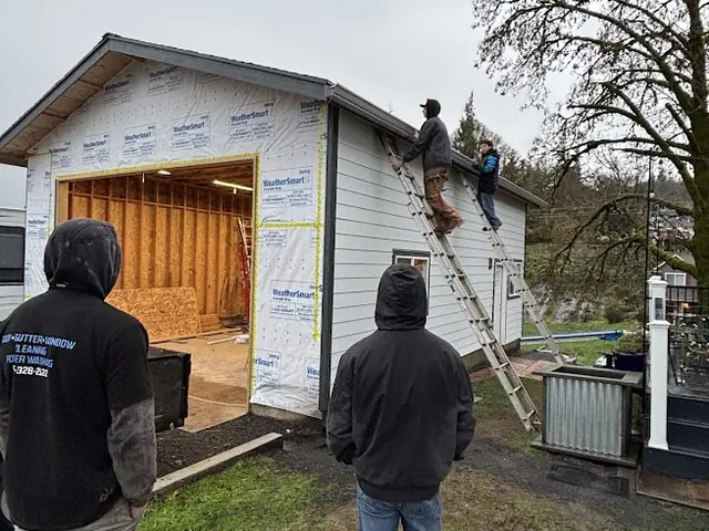 Crew members on ladders at the eave of a new garage wrapped in house wrap in Columbia City, with two workers watching from the lawn