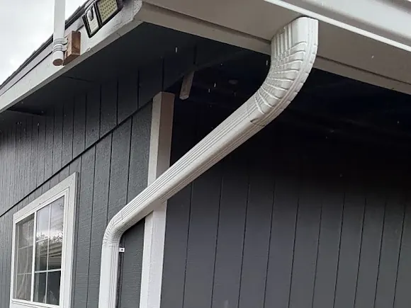 White downspout with offset elbows running from the gutter outlet under the eave and down a gray-sided home