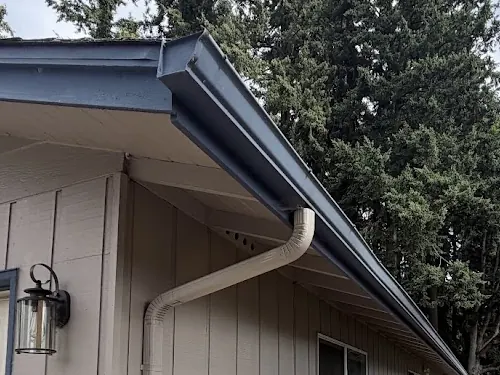 Gray seamless gutter with a matching downspout and offset elbow running down the side of a tan garage under tall evergreens
