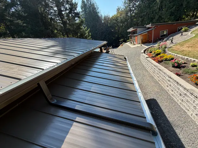 View from above of a gutter along the edge of a dark metal roof in St. Helens, with a gravel drive and flower beds below
