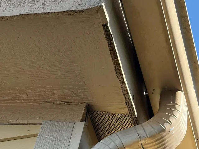 Close-up under a Scappoose eave showing the back of a tan gutter and a downspout elbow connected at the outlet