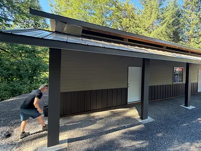 AGS crew member at work beside a new metal-roofed shop building under tall firs near St. Helens
