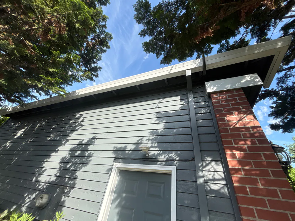 Looking up at a light-colored gutter along a garage eave under evergreen branches, with a gray downspout beside a brick column