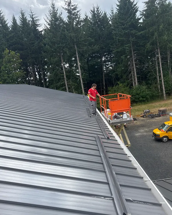 American Gutter Service installer on a lift hanging a new seamless gutter along a metal shop roof with tall fir trees behind