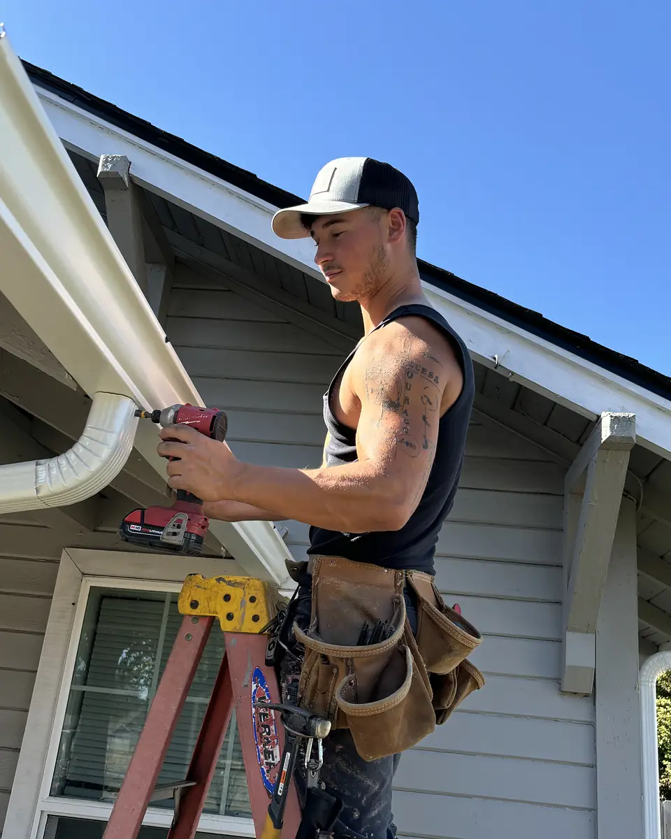 American Gutter Service technician on a ladder driving a screw into a white gutter beside a downspout elbow