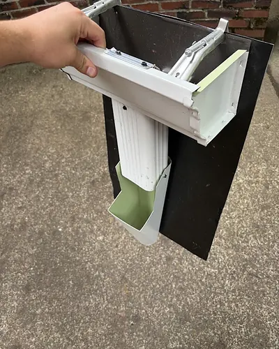 Cut section of white K-style seamless gutter with a downspout outlet attached, held up to show the gutter profile