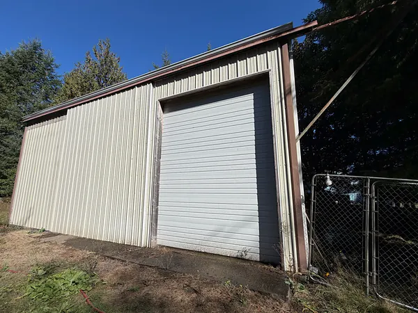 Metal shop building with a tall roll-up door and a seamless gutter along the eave on a wooded Clatskanie property