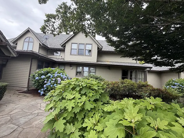 Large tan two-story Oregon home with new gutters along multiple rooflines behind a landscaped yard with hydrangeas