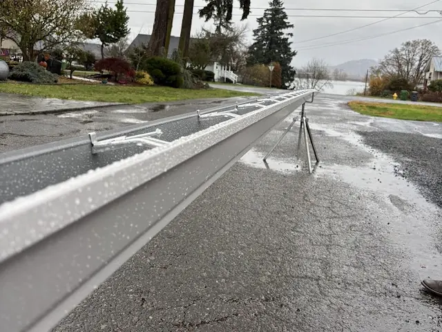 A long seamless gutter run resting on stands along a wet residential street in Columbia City