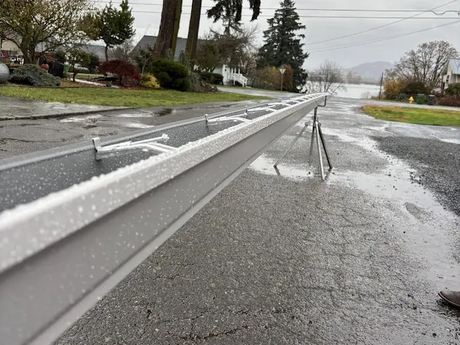 A long seamless gutter run resting on stands along a wet residential street in Columbia City