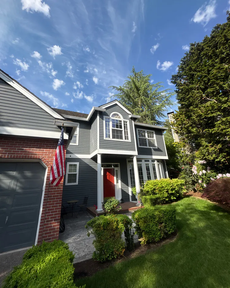 Front of a dark gray two-story house with white trim, a red door, a brick-faced garage, and roof edges at several heights