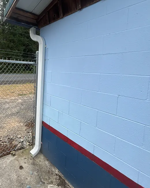 White downspout running down the painted block wall of a shop building in Oregon, beside a chain-link fence and tall firs