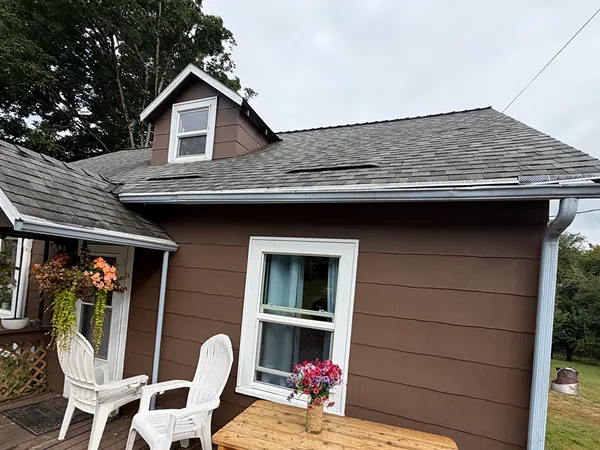 Brown Clatskanie farmhouse with a dormer, white gutters and a white downspout beside a wood deck with Adirondack chairs