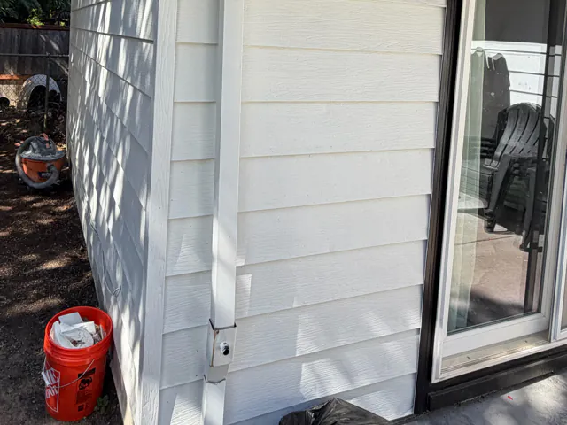 White downspout with a cleanout fitting near the bottom, running down white lap siding beside a sliding glass door