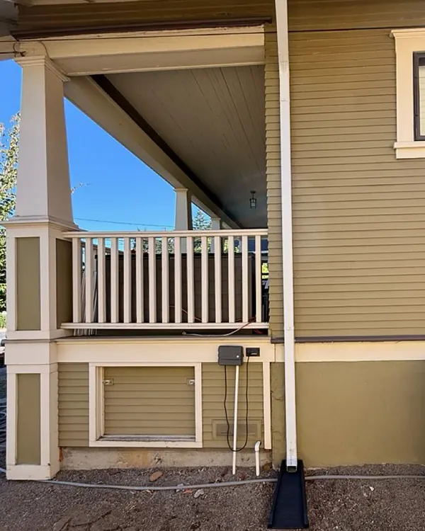 White downspout running down an olive-green Portland craftsman beside the front porch and ending on a black splash block
