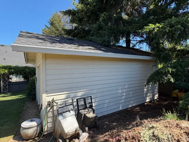 White gutter along the eave of a cream-sided detached garage in Warren under large fir trees