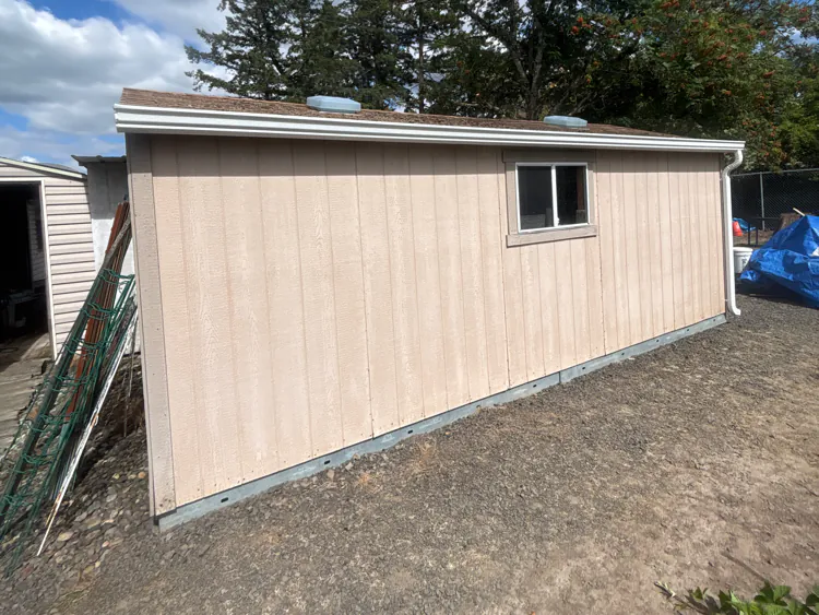 White gutter along the eave of a tan detached shed, with a white downspout at the far corner and trees behind
