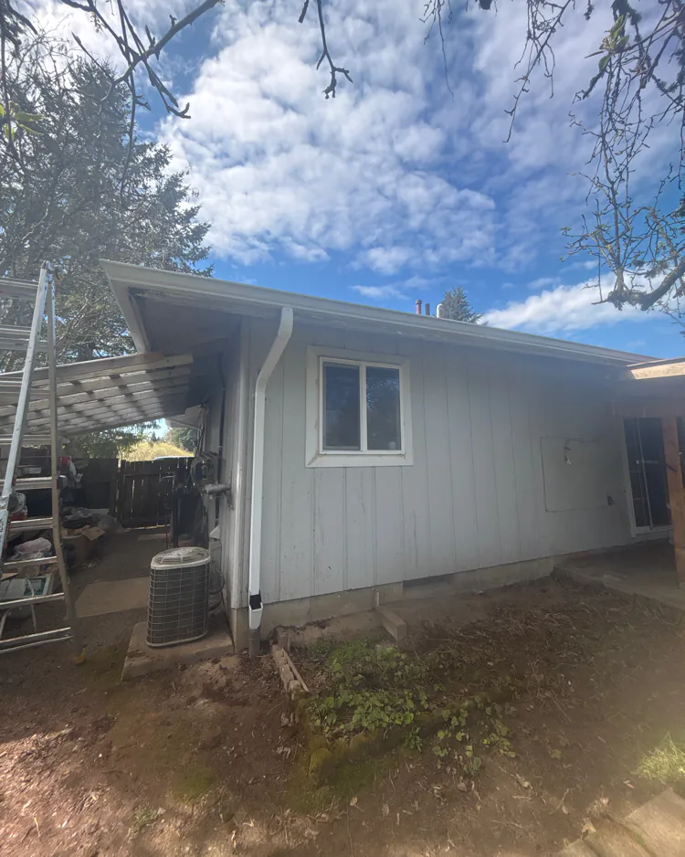 White gutter along the roof edge of a light gray single-story house, with a white downspout at the corner beside an air-conditioning unit and a ladder