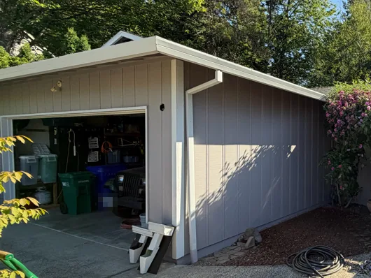White gutter running along a gray garage eave and around the corner, with a white downspout down the side wall and spare downspout pieces on the ground