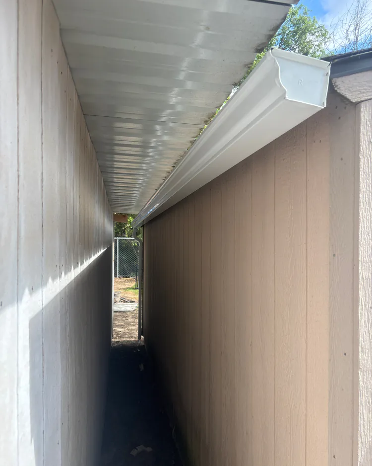 White K-style gutter with an end cap running along a tan shed wall in a narrow side passage, with a downspout at the far end