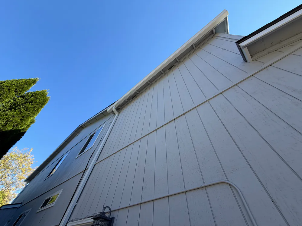 Looking up a tall gray wall at a white gutter running along a high eave, with a white downspout down the corner