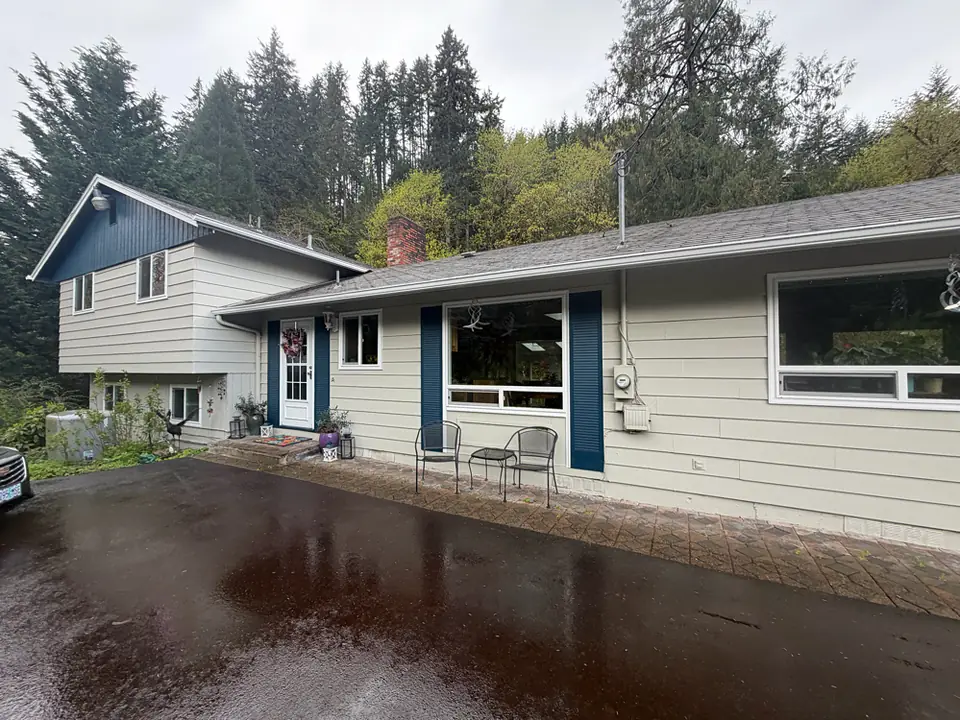 Split-level Oregon home with white gutters and downspouts, a rain-soaked driveway, and a hillside of evergreen trees behind