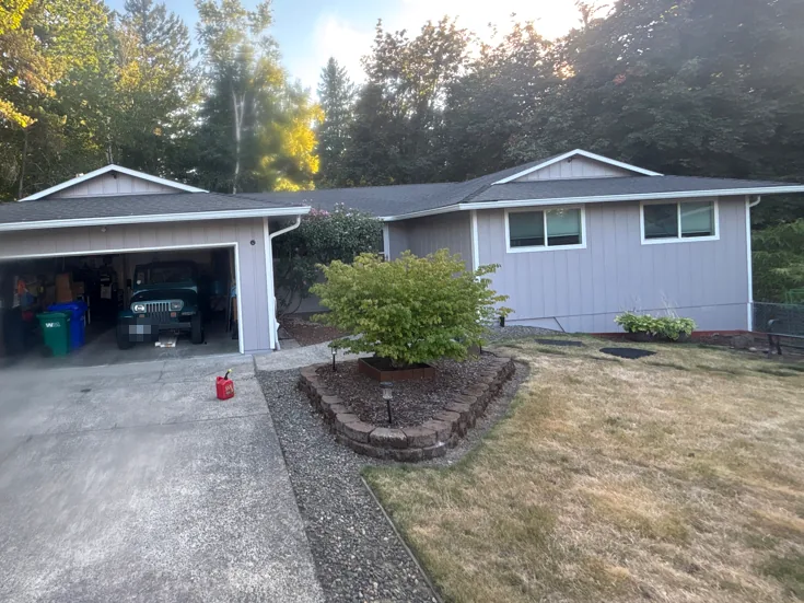 Single-story gray home with white gutters along the garage and main roof eaves, a white downspout at the garage corner, and tall trees behind the house