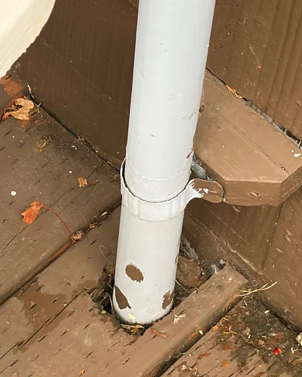 Old, paint-spotted round downspout running through a rotting wood deck, an example of a downspout that needs replacing