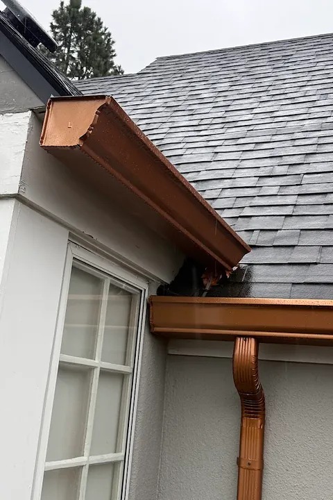 Copper-colored K-style gutter and downspout at an inside corner of a white stucco home