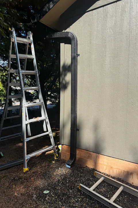 Dark bronze downspout running down gray panel siding to the ground, with a ladder beside it