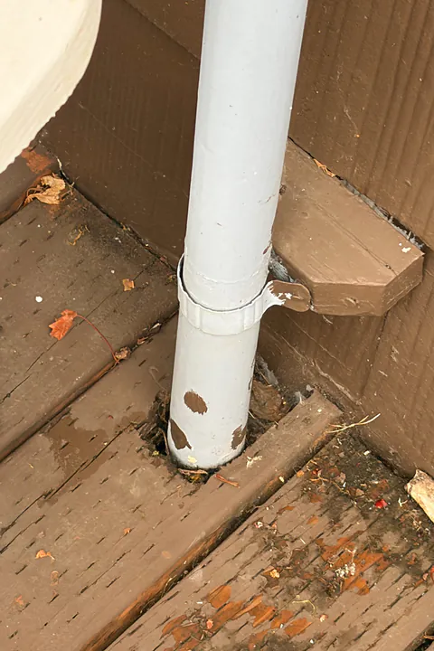 White downspout passing down through an opening in deck boards with rust stains around it