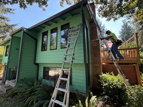 Installer on a ladder fitting a dark gutter beside the brick chimney of a green two-story home
