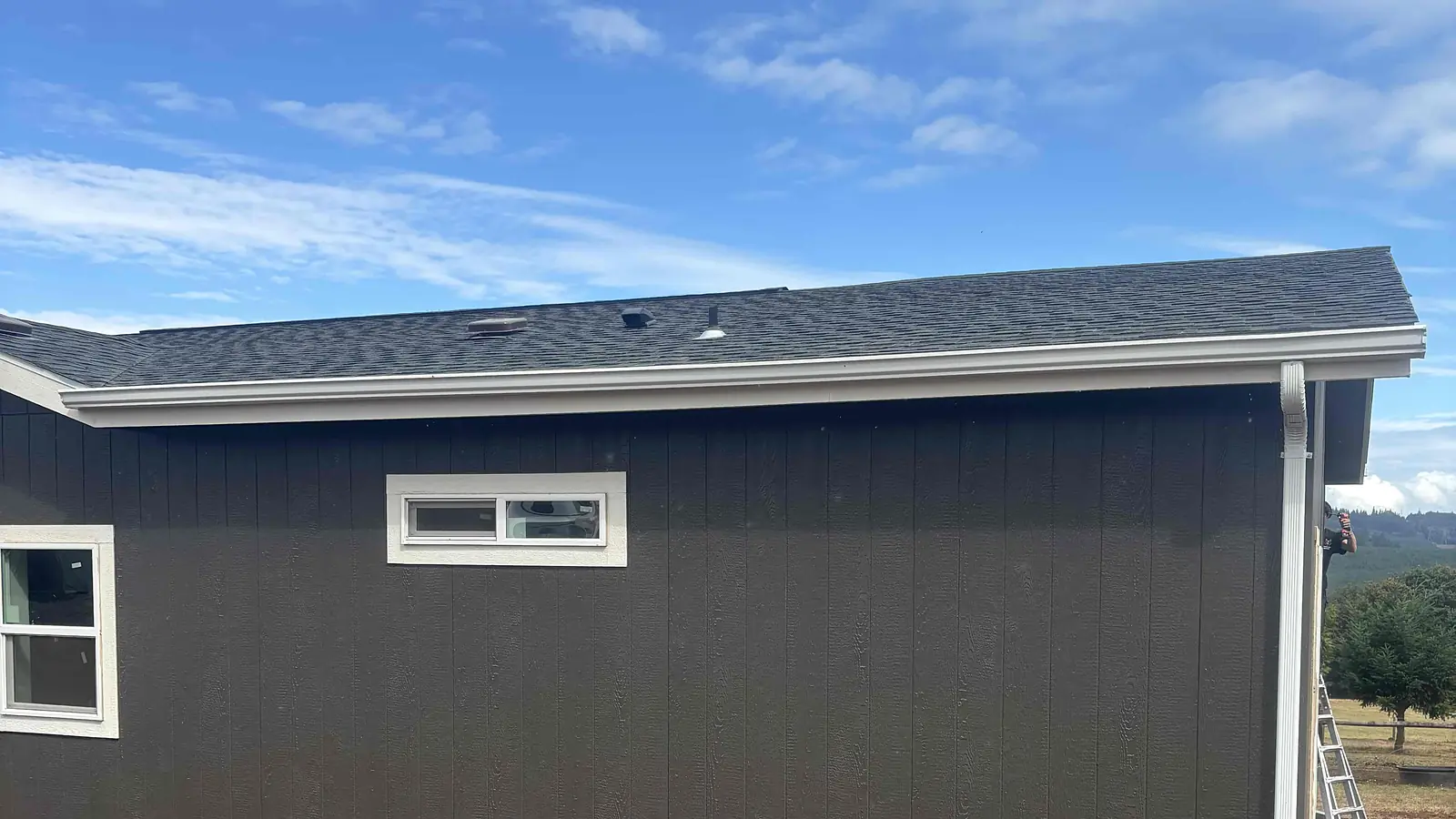 Single-story home in Deer Island, Oregon with new white seamless gutters and a corner downspout under a cloudy sky