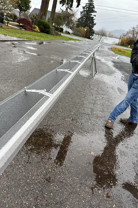 Long white seamless gutter with hidden hangers set inside, lying on a wet street before hanging