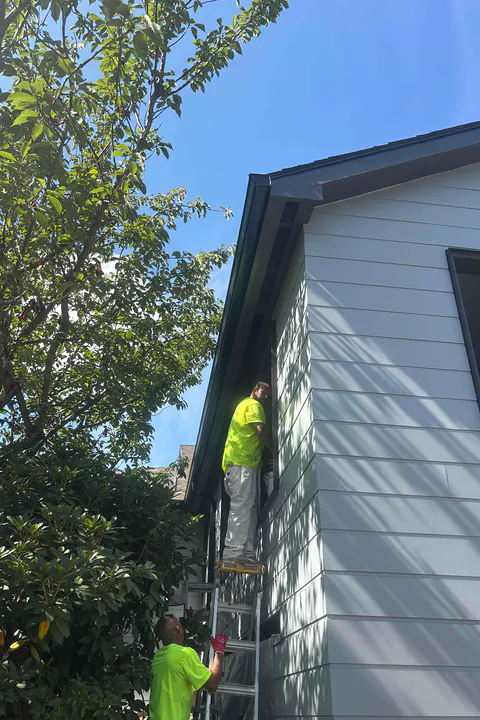 Two installers in high-visibility shirts on ladders at the corner of a light gray building