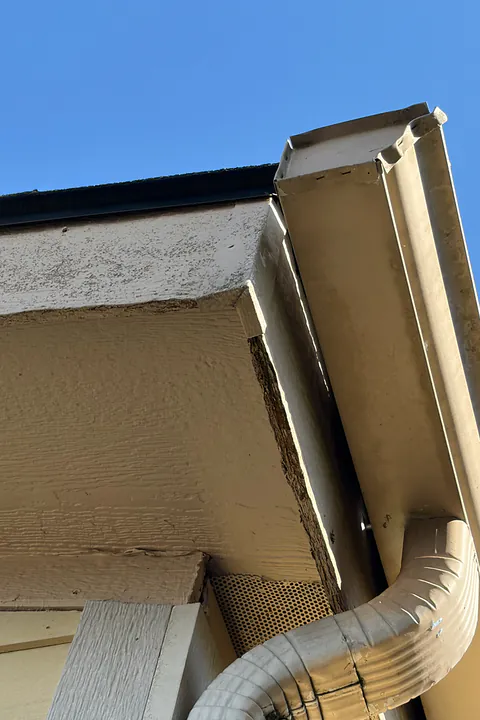 Tan gutter corner and downspout elbow where the fascia trim behind it is cracked and weathered