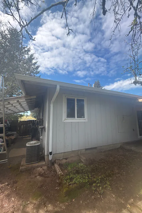 Corner of a light gray ranch home with a white gutter and downspout beside a patio cover in Tigard, Oregon