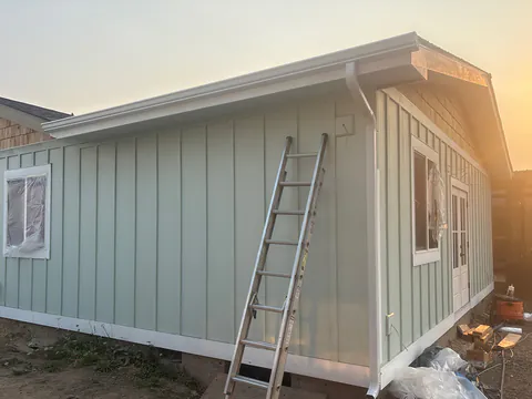 New sage board-and-batten addition with a white gutter and downspout and a ladder against the wall