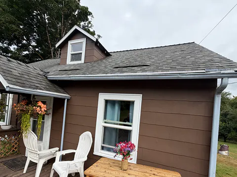 Brown farmhouse with a dormer, white gutters and a white downspout beside a deck with chairs