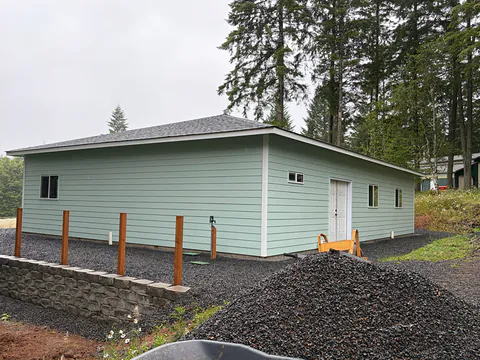Light green hip-roof building on a gravel lot with white gutters, new fence posts and tall firs