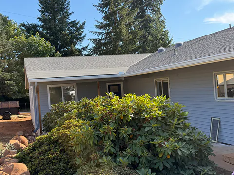 Light blue ranch home with white seamless gutters, a large rhododendron and tall firs behind